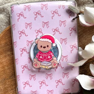 Könnte beinhalten: Eine transparente Handyhülle mit einem rosa Schleifenmuster. Ein Pop-Socket zeigt einen Teddybären mit Weihnachtsmütze und Weihnachtspullover, der auf Büchern sitzt. Der Hintergrund ist hellrosa.