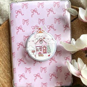 Op de afbeelding: Een doorzichtige beschermhoes voor een e-reader met een roze strikpatroon. Een decoratieve popsocket toont een kerstscène met een huis, boom en hond. De hoes is ontworpen om de e-reader te beschermen.