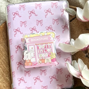 Könnte beinhalten: Eine transparente Tablet-Hülle mit rosa Schleifenmuster, die einen rosa-weißen Bookstore-Pop-Socket hält. Der Pop-Socket zeigt einen Geist, Bücher und das Wort "BOOKSTORE". Ein Schild "Sale 30%" ist sichtbar.