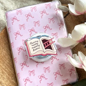 Könnte beinhalten: Eine rosa Tablet-Hülle mit einem sich wiederholenden Muster aus rosa Schleifen. Ein Pop-Socket mit einem offenen Buchdesign mit der Aufschrift "Leaving reality entering fiction" ist an der Hülle befestigt. Das Buchdesign zeigt eine Leiter und einen Sternenhimmel.