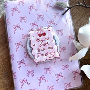 Puede incluir: Una funda protectora transparente para lector de libros electrónicos con un patrón de lazos rosas. Una pegatina decorativa con el texto "Buy me books & tell me I'm pretty" está adherida, con un diseño de cuadros vichy e ilustraciones de cerezas.