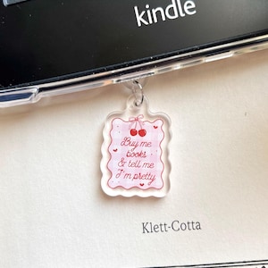 Op de afbeelding: Een heldere acryl bedel met een roze ruitpatroon en de tekst "Buy me books & tell me I'm pretty". De bedel heeft een kersendesign en is bevestigd aan een Kindle.