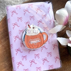 Puede incluir: Una funda transparente para teléfono con un patrón de lazos rosas sobre fondo lila claro. Un adhesivo circular naranja y blanco con un café con leche de calabaza y la frase "Bookish Spice Latte".