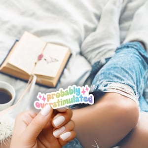 May include: A sticker with the text "probably overstimulated" in rainbow colors. The sticker is held by a person wearing blue jeans and white socks.