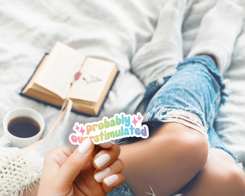 May include: A sticker with the text "probably overstimulated" in rainbow colors. The sticker is held by a person wearing blue jeans and white socks.