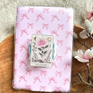 May include: A clear protective case with a pink bow pattern, holding a tarot card with a rose and the text "THE READER". The case is on a wooden tray with a flower.