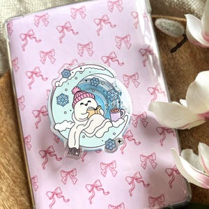 May include: A clear phone case with a pink bow pattern. A circular pop socket features a snowman reading a book on a crescent moon, with snowflakes and a mug of hot cocoa. The snowman wears a pink hat and scarf.