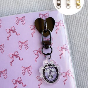 Puede incluir: Un cierre negro en forma de corazón sujeto a un cuaderno morado claro con un patrón de lazos rosas. Un dije con el texto "The Villains Book Club" y una ilustración de un pájaro cuelga del cierre. Tres tiradores de cremallera en forma de corazón en negro, plata y oro.