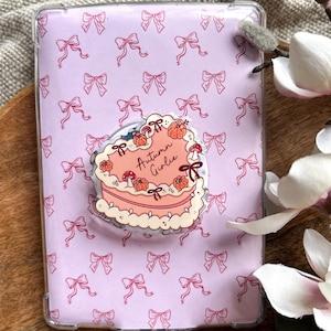 Può includere: Una custodia trasparente per telefono con sfondo lilla chiaro decorato con fiocchi rosa. Un adesivo a forma di torta rosa con zucche e la scritta "Autumn Girlie" è attaccato. La custodia è su una superficie di legno.