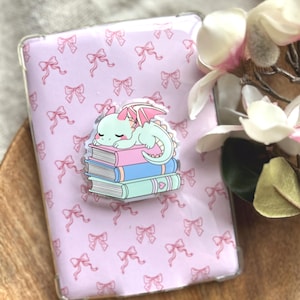 Könnte beinhalten: Eine rechteckige Handyhülle mit hellrosa Hintergrund und einem wiederkehrenden Muster aus kleinen rosa Schleifen. In der Mitte befindet sich ein Acryl-Anhänger, der einen kleinen, hellgrünen Drachen zeigt, der auf drei Büchern schläft.