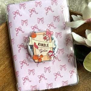 Op de afbeelding: Een rechthoekige e-readerhoes met een lichtroze achtergrond en een herhalend patroon van kleine rode strikjes. In het midden is een decoratieve acrylgreep bevestigd met de tekst Kindle Girlie, pompoenen, paddenstoelen en herfstbladeren.