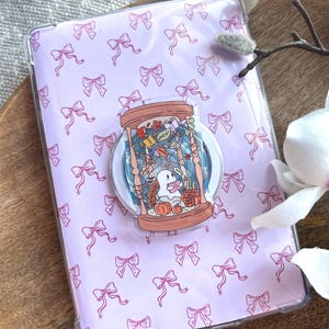 Op de afbeelding: Een rechthoekige tablethoes met een lichtpaarse achtergrond met een herhalend patroon van kleine roze strikjes. Een ronde telefoonhouder is in het midden bevestigd, met een illustratie van een geest in een zandloper met boeken en pompoenen.