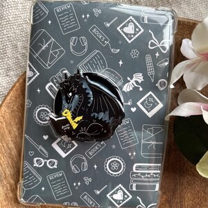 Puede incluir: Una funda protectora transparente para lector electrónico con fondo gris oscuro y dibujos lineales blancos de libros, auriculares y formularios. Un soporte negro con forma de dragón muestra a un dragón leyendo un libro titulado Fourth Wing.