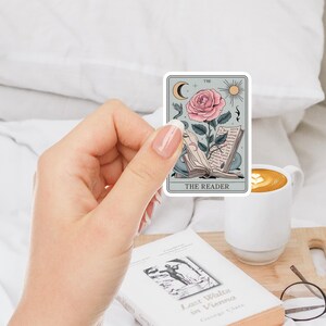 May include: A sticker featuring a tarot card illustration of The Reader. The card depicts a pink rose with a stem and leaves growing from an open book. The card has a light blue background with a crescent moon, sun, and musical notes.