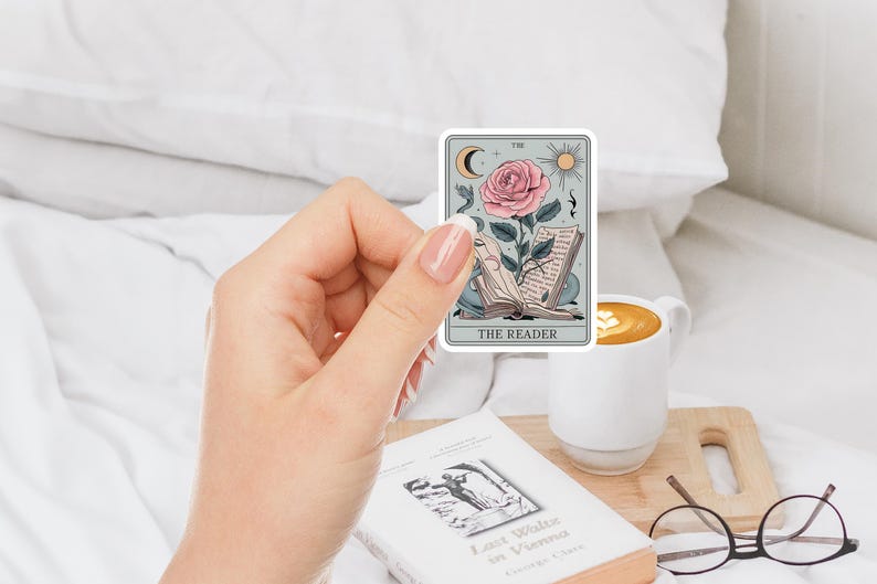 May include: A sticker featuring a tarot card illustration of The Reader. The card depicts a pink rose with a stem and leaves growing from an open book. The card has a light blue background with a crescent moon, sun, and musical notes.