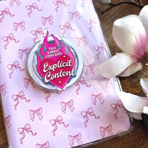 Könnte beinhalten: Eine transparente Handyhülle mit einem rosa Schleifenmuster und einem Pop-Socket. Der Pop-Socket hat ein rosa Herzdesign mit den Worten "THIS KINDLE CONTAINS Explicit Content" in einem flammenartigen Rahmen.
