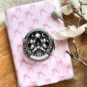 May include: A clear phone case with a pink bow pattern, featuring a black and white pop socket. The pop socket has a mountain and hooded figure design with the text "Suriel Tea Co."