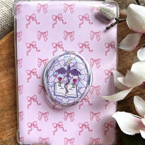 Op de afbeelding: Een doorzichtige beschermhoes voor een e-reader met een lichtroze strikpatroon. Een decoratieve popsocket met een glas-in-lood ontwerp met een draak, rozen en een boek is aan de hoes bevestigd.
