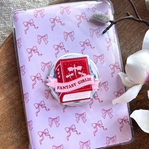 Puede incluir: Una funda transparente para teléfono con un patrón de lazos de color lila claro. Un pop socket con un diseño de libro rojo y las palabras "FANTASY GIRLIE" está adherido a la funda. La funda está sobre una bandeja de madera con una flor.