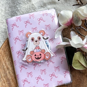 Puede incluir: Una funda de teléfono transparente con fondo morado claro y un patrón repetitivo de pequeños lazos rosas. En el centro hay un dije de acrílico de un oso marrón con un disfraz de fantasma blanco que sostiene un cubo de calabaza rosa.