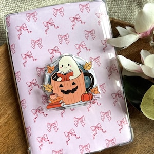 Op de afbeelding: Een doorzichtige telefoonhoes met een roze strikpatroon. Een Halloween-thema popsocket toont een spook dat leest in een pompoenbeker met herfstbladeren, een stapel boeken en een kleine pompoen.