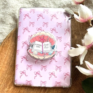 May include: A pink tablet case with a repeating bow pattern and a seashell-shaped pop socket. The pop socket features the text "Summer Girl Book Club" and a starfish illustration.