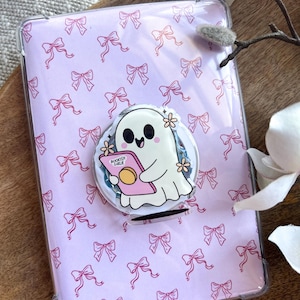 Könnte beinhalten: Eine transparente Handyhülle mit einem rosa Schleifenmuster. Ein weißer, geisterförmiger Handyhalter ist angebracht, der ein rosa Buch mit dem Text "BOOKISH GIRL" hält. Der Halter hat ein Blumenmuster, auf einer Holzoberfläche.
