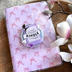 Könnte beinhalten: Eine transparente Kindle-Hülle mit einem hellvioletten Schleifenmuster. Ein kreisförmiger Pop-Socket zeigt ein Buchdesign mit dem Text "a reader without their Kindle is a tragedy". Die Hülle liegt auf einer Holzoberfläche mit einem Magnolienzweig.