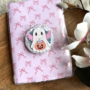 Könnte beinhalten: Eine transparente Schutzhülle für einen E-Reader mit einem rosa Schleifenmuster. Ein kreisförmiger Pop-Socket mit einem Cartoon-Geist, der einen Kürbis hält, ist angebracht. Der Geist hat rosa Flügel und Sterne.