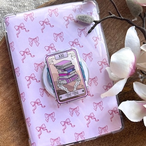 Op de afbeelding: Een doorzichtige telefoonhoes met een lichtroze strikpatroon. Een telefoonhouder met tarotthema toont een skelet dat boeken vasthoudt. De kaart leest "XXI" en "THE TBR".