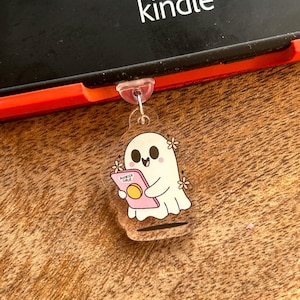 Può includere: Un portachiavi in acrilico trasparente con un fantasma dei cartoni animati che tiene in mano un libro rosa con la scritta "Bookish Girlie". Il fantasma ha una faccia sorridente e piccoli fiori. Il portachiavi è attaccato a un Kindle nero.