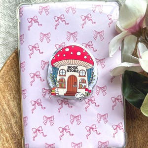 Könnte beinhalten: Eine rechteckige Handyhülle mit hellrosa Hintergrund und einem sich wiederholenden Muster aus kleinen rosa Schleifen. Ein runder Aufkleber ist mittig auf der Hülle angebracht und zeigt einen roten pilzförmigen Buchladen mit dem Text BOOK STORE.