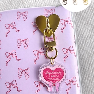 Puede incluir: Un tirador de cremallera dorado en forma de corazón con un mosquetón, sujeto a un charm acrílico transparente. El charm es rosa con el texto "Buy me books & tell me I'm pretty" e ilustraciones de lazos. Fondo lila con patrones de lazos rosas.