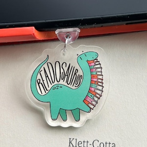 Op de afbeelding: Een heldere acryl sleutelhanger met een turquoise dinosaurus met een ruggengraat van kleurrijke boeken. De sleutelhanger leest "READOSAURUS". De achtergrond bevat een Kindle-apparaat.