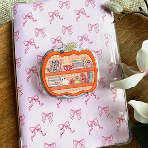 Könnte beinhalten: Eine transparente Schutzhülle für einen E-Reader mit einem rosa Schleifenmuster. Ein kürbisförmiger Pop-Socket ist angebracht, der ein Bücherregal-Design mit Büchern, einem Geist und anderen dekorativen Elementen aufweist.