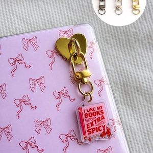 Puede incluir: Un tirador de cremallera dorado en forma de corazón, sujeto a una funda transparente para teléfono con un estampado de lazos rosas. Un pequeño dije con el texto "I LIKE MY BOOKS EXTRA SPICY" cuelga del tirador de la cremallera. Se muestran tres tiradores de cremallera adicionales en un círculo.
