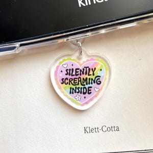 May include: A clear heart-shaped charm with the text "SILENTLY SCREAMING INSIDE" in black, surrounded by a rainbow design and a scalloped edge. The charm is attached to a silver-colored metal ring. A black Kindle device is visible in the background.