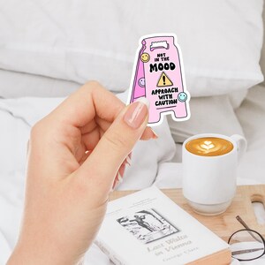Op de afbeelding: Roze en witte waarschuwingsbord sticker met de tekst "Niet in de stemming. Benader met voorzichtigheid." en een waarschuwingssymbool. De sticker heeft een cartoon smiley op elke hoek.