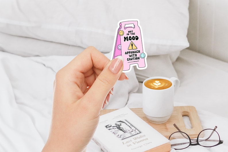 Op de afbeelding: Roze en witte waarschuwingsbord sticker met de tekst "Niet in de stemming. Benader met voorzichtigheid." en een waarschuwingssymbool. De sticker heeft een cartoon smiley op elke hoek.