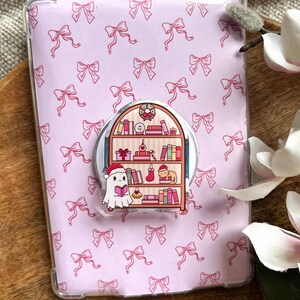 Könnte beinhalten: Eine hellrosa Tablet-Hülle mit einem sich wiederholenden Muster aus rosa Schleifen. Ein Pop-up-Griff zeigt einen Cartoon-Geist mit Weihnachtsmütze, der ein Buch vor einem Bücherregal mit Weihnachtsdekorationen liest.