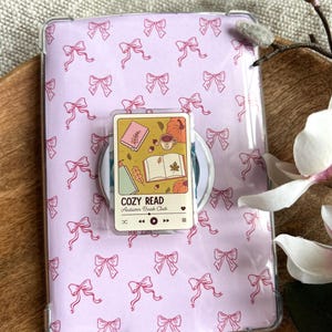 Op de afbeelding: Een doorzichtige tablethoes met een herhalend patroon van roze strikken op een lichtpaarse achtergrond. In het midden is een rechthoekige telefoonhouder bevestigd met een afbeelding van een open boek, een mok en pompoenen, gelabeld COZY READ Autumn Book Club.