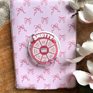 Könnte beinhalten: Eine rosa Tablet-Hülle mit einem sich wiederholenden Schleifenmuster und einem "Mood Wheel" Pop-Socket. Der Pop-Socket trägt das Wort "SMUTTY" und Sätze wie "Slow Burn" und "Touch Her & Die".