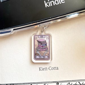 Può includere: Un ciondolo in acrilico trasparente con un design di carta dei tarocchi che mostra una pila di libri tenuta da mani scheletriche. La carta recita "THE TBR" e "XXI". Il ciondolo è appeso vicino a un e-reader Kindle. Il testo "Klett-Cotta" è sotto.