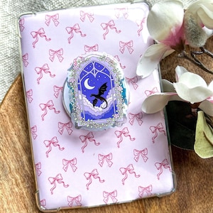 Puede incluir: Una funda transparente para teléfono con un patrón repetitivo de pequeños lazos rosas sobre un fondo morado claro, con un soporte circular para teléfono en el centro que muestra una ilustración de ventana gótica con un dragón negro, luna y nubes.
