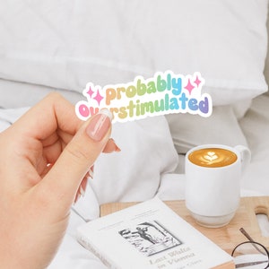 May include: A rainbow sticker with the text "Probably Overstimulated" with pink glitter stars on either side. The sticker is being held by a hand.
