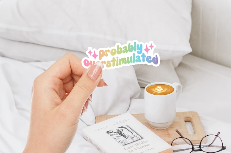May include: A rainbow sticker with the text "Probably Overstimulated" with pink glitter stars on either side. The sticker is being held by a hand.