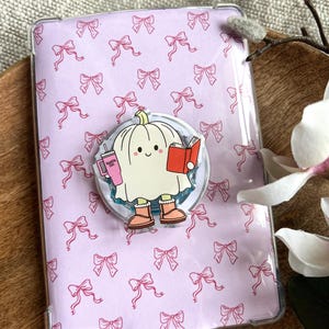Puede incluir: Una funda transparente para tableta con un fondo lila claro cubierto de lazos rosas. Un pop socket con un fantasma de dibujos animados leyendo un libro y sosteniendo una bebida está adjunto. El fantasma lleva botas naranjas.