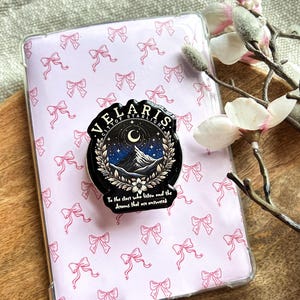 Könnte beinhalten: Eine Handyhülle mit einem rosa Schleifenmuster und einer schwarzen Anstecknadel. Die Anstecknadel zeigt den Text "VELARIS CITY OF STARLIGHT" um ein Berg- und Monddesign, mit dem Zitat "To the stars who listen and the dreams that are answered."