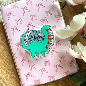 Può includere: Una custodia trasparente per tablet con un motivo a fiocchi rosa. Un adesivo a forma di dinosauro turchese con la scritta "READOSAURUS" e una pila di libri è attaccato. La custodia è su una superficie di legno.