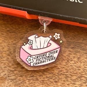 Puede incluir: Un llavero acrílico transparente con un diseño de caja de pañuelos rosa. La caja de pañuelos tiene pañuelos blancos y el texto "Crying Over Fictional Characters". El llavero tiene un anillo de metal plateado y un lazo de plástico transparente.